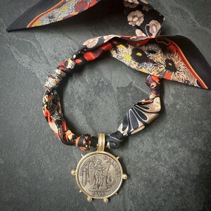 Puede incluir: Un collar con un colgante de moneda vintage en un marco dorado, suspendido de una bufanda de seda estampada. La bufanda presenta un diseño floral en negro, naranja y crema. La moneda tiene un diseño grabado y el año 1793.
