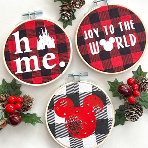 May include: Three round Christmas ornaments in wooden hoops. The ornaments feature red and black buffalo plaid fabric with white text and graphics. One says "home." with a castle graphic, another says "Joy to the World" with a Mickey Mouse head, and the third has a Mickey Mouse head with a snowflake.