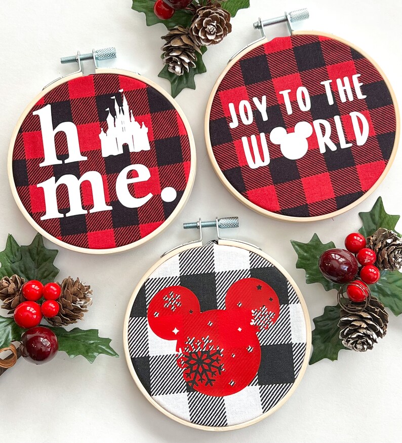 May include: Three round Christmas ornaments in wooden hoops. The ornaments feature red and black buffalo plaid fabric with white text and graphics. One says "home." with a castle graphic, another says "Joy to the World" with a Mickey Mouse head, and the third has a Mickey Mouse head with a snowflake.