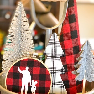 May include: A decorative arrangement featuring a silver glittered Christmas tree, a red and black plaid fabric hoop with a silhouette design, and various wooden Christmas tree decorations. The items are displayed in a galvanized metal container.