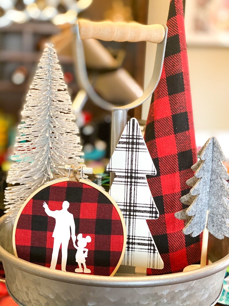 May include: A decorative arrangement featuring a silver glittered Christmas tree, a red and black plaid fabric hoop with a silhouette design, and various wooden Christmas tree decorations. The items are displayed in a galvanized metal container.
