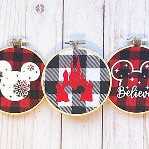 May include: Three round embroidery hoops with a red and black buffalo plaid pattern. Each hoop features a different design: a Mickey Mouse head with snowflakes, a red castle, and a Mickey head with the word "Believe".