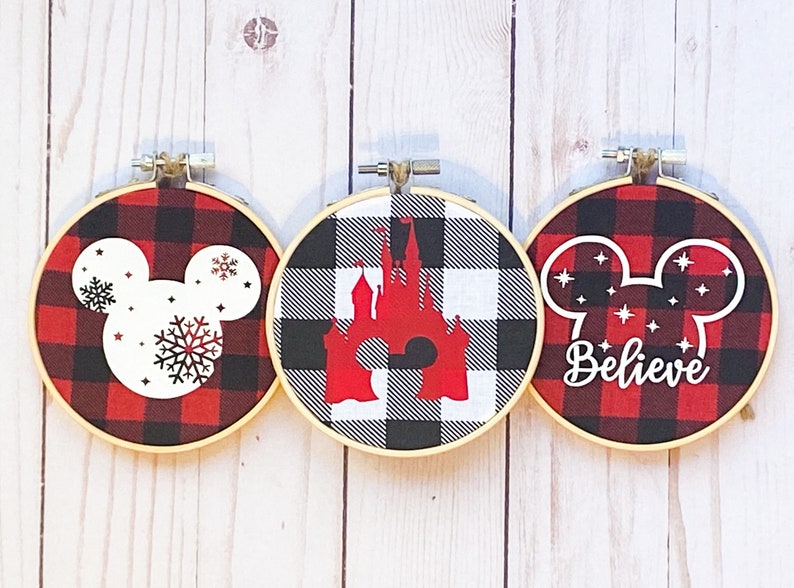 May include: Three round embroidery hoops with a red and black buffalo plaid pattern. Each hoop features a different design: a Mickey Mouse head with snowflakes, a red castle, and a Mickey head with the word "Believe".