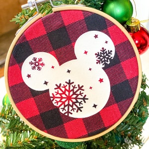 May include: A round Christmas ornament featuring a red and black buffalo plaid pattern. The ornament has a white Mickey Mouse head shape with snowflake and star designs. The ornament is held in a wooden embroidery hoop.