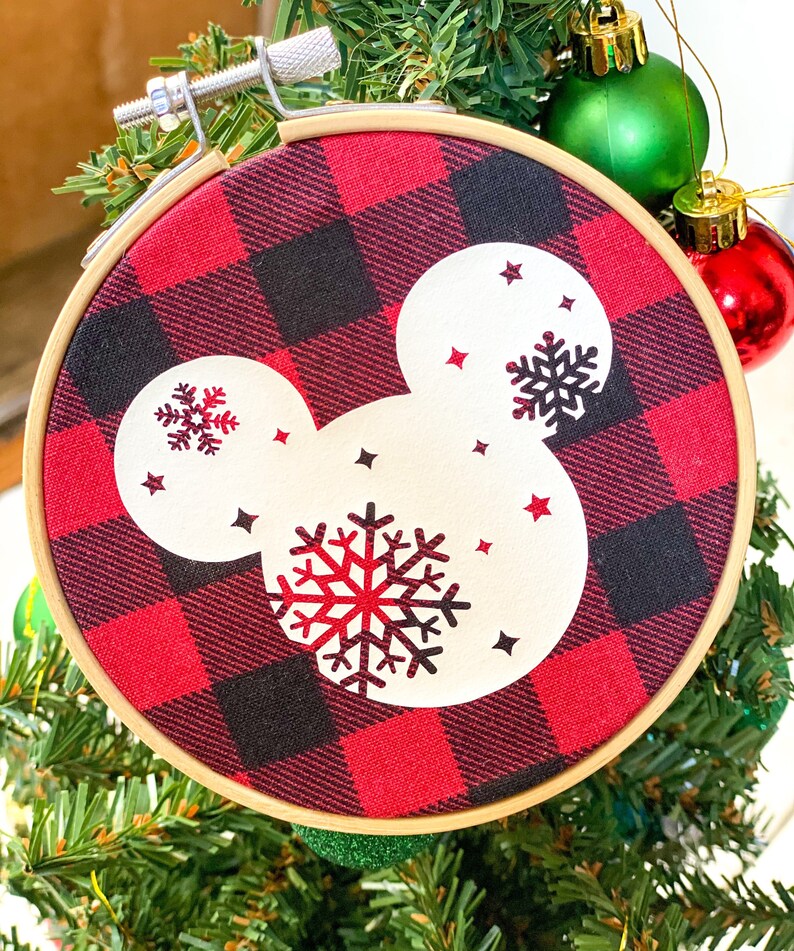 May include: A round Christmas ornament featuring a red and black buffalo plaid pattern. The ornament has a white Mickey Mouse head shape with snowflake and star designs. The ornament is held in a wooden embroidery hoop.