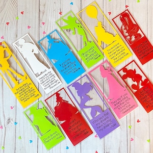 May include: A set of 12 colorful bookmarks featuring Disney characters and quotes. Each bookmark is made of clear plastic with a colorful silhouette of a Disney character and a quote from the character. The bookmarks are arranged in a row on a wooden surface.