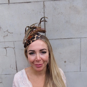 Puede incluir: Tocado decorativo con estampado de leopardo y plumas marrones. El tocado se lleva en la cabeza. El fondo es una pared de ladrillos gris claro. El tocado es un accesorio de moda.