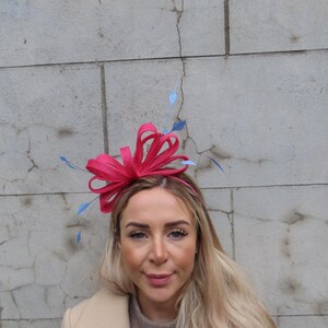 May include: A pink and blue headband with a large bow and feathers. The headband is worn by a person with long blonde hair.