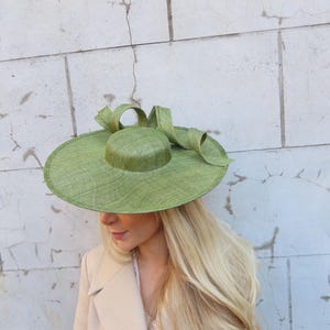 May include: A wide-brimmed, light green hat with a rounded crown and a large bow. The hat is made of woven material and is worn with a beige blazer. The hat's brim is approximately 15 inches in diameter.