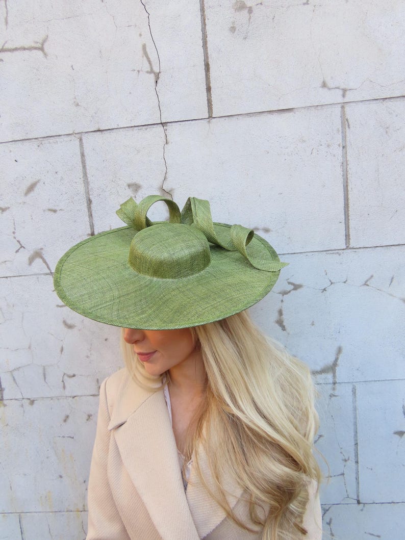 May include: A wide-brimmed, light green hat with a rounded crown and a large bow. The hat is made of woven material and is worn with a beige blazer. The hat's brim is approximately 15 inches in diameter.