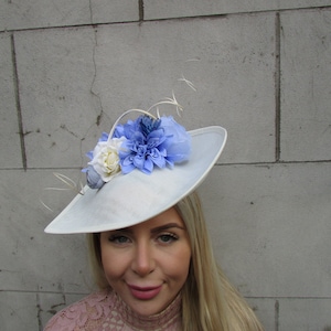 Può includere: Cappello azzurro e bianco con decorazioni floreali. Il cappello presenta un'ampia tesa ed è decorato con fiori di stoffa blu e bianchi e delicati rametti. Questo elegante cappello è adatto per occasioni speciali.