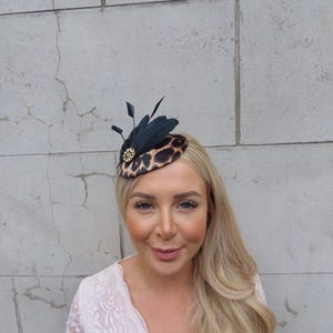 May include: A small, round, leopard print hat with a black feather accent. The hat is adorned with a gold-colored jewel.