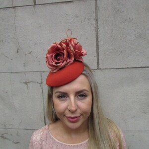 May include: A red felt hat with a coral rose flower embellishment. The hat is a small, round shape and is worn on the side of the head.