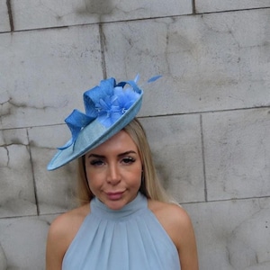 May include: A light blue fascinator hat featuring a wide, angled brim, a textured fabric base, and decorative elements including layered fabric loops and soft, sheer floral accents positioned on the top side of the headpiece.