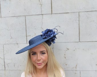 May include: A navy blue hat with a wide brim and a large flower accent. The hat is worn at an angle, and the flower is positioned on the side. The hat is a stylish accessory, suitable for special occasions.