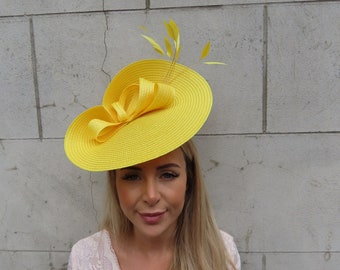 May include: A bright yellow, wide-brimmed hat with a large bow and feather accents. The hat is made of a textured material and is designed for special occasions. The hat is a vibrant accessory.