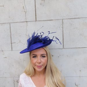 May include: A royal blue hat with a wide brim and a cluster of dark blue feathers. The hat is a vibrant accessory, perfect for special occasions. The hat is set against a textured, light-colored wall.