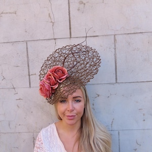May include: A brown, woven headpiece with a circular design, adorned with several pink roses. The headpiece is worn on the head. The background is a light-colored brick wall. This headpiece is suitable for special occasions.