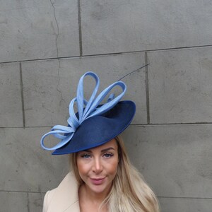 May include: A navy blue fascinator hat featuring a wide, rounded base and a series of light blue, looped, ribbon-like structures extending upward from the side. The hat has a smooth, structured texture and includes a single thin, dark quill accent.
