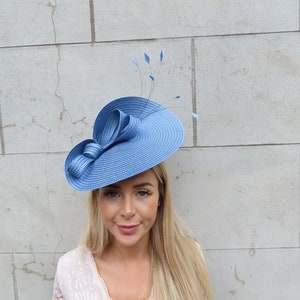 May include: A blue fascinator hat featuring a circular base with a textured, woven appearance. The hat is adorned with a series of looped ribbons on one side and several thin, delicate stems topped with small, pointed blue feather accents.