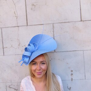 May include: A light blue hat with a wide brim and a decorative bow. The hat is made of woven material and has a few long, thin accents. The hat is worn by a person with long blonde hair and a white lace top.