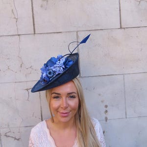 May include: A navy blue hat with a wide brim, adorned with various shades of blue fabric flowers and a single blue feather. The hat is a formal accessory, suitable for special occasions.
