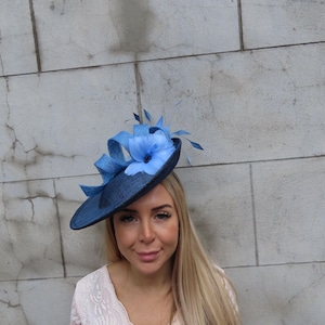 May include: A navy blue hat with a wide brim, adorned with a light blue flower and matching ribbon. The hat features delicate blue feathers. This elegant headwear is suitable for special occasions.
