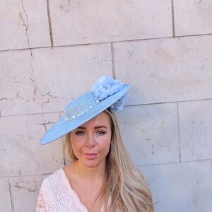 May include: A light blue hat with a wide brim, adorned with pearls and a matching fabric flower. The hat is a pastel blue color and appears to be made of woven material. A stylish accessory for special occasions.