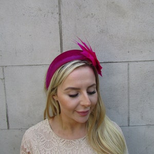 May include: A fuchsia velvet headband with a matching feather and flower accent. The headband is worn by a person with long blonde hair.