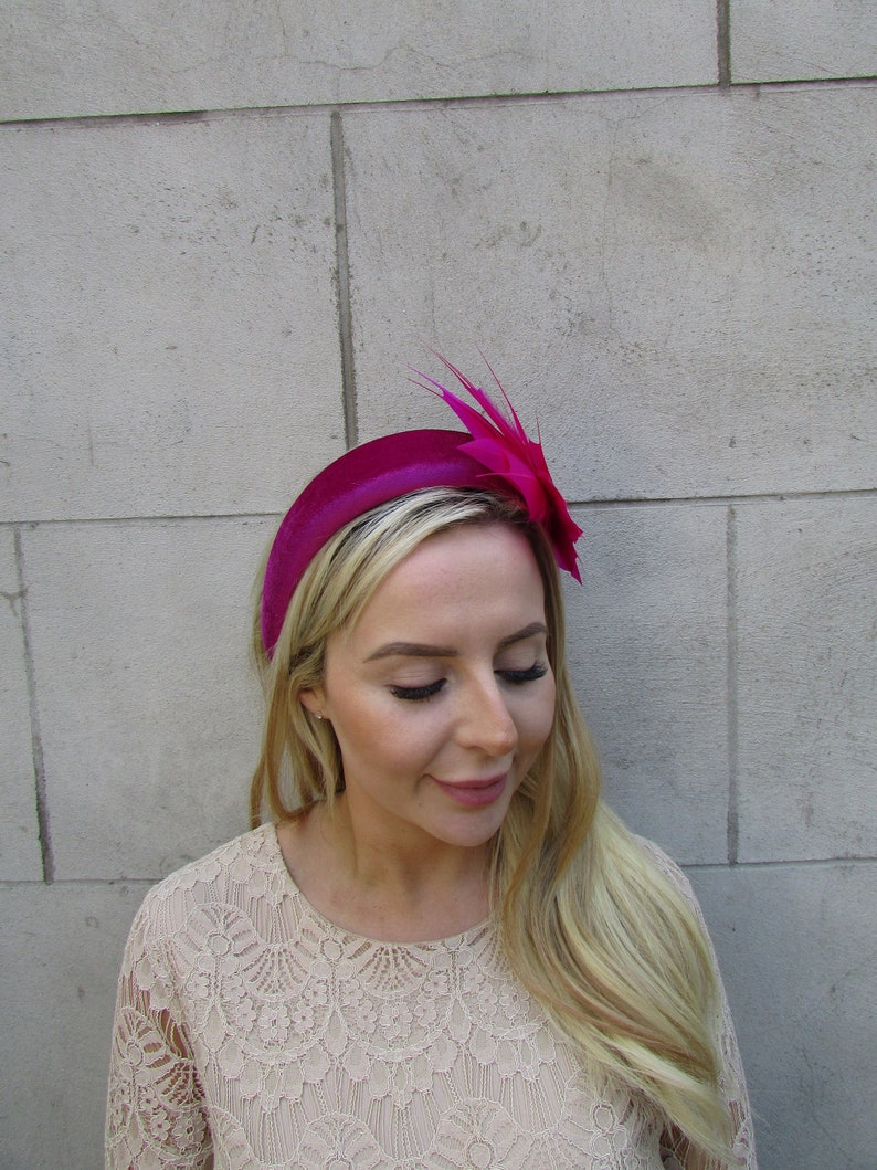May include: A fuchsia velvet headband with a matching feather and flower accent. The headband is worn by a person with long blonde hair.