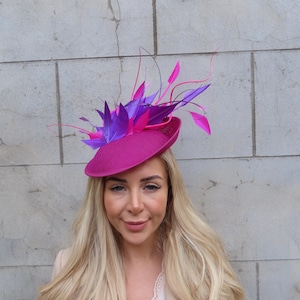 Puede incluir: Un pequeño sombrero redondo de color fucsia con un adorno de plumas moradas y rosas.