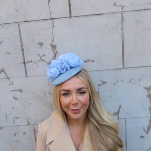 Könnte beinhalten: Hellblauer Fascinator mit drei großen, passenden Stoffblumen. Die Person trägt einen beigen Mantel. Der Hintergrund ist eine hellgraue Steinmauer.