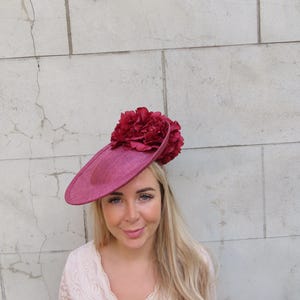 Könnte beinhalten: Ein magentafarbener Fascinator mit einer breiten, flachen, kreisförmigen Krempe und einem Büschel dunkelroter Kunstblumen auf der Oberseite. Der Hut besteht aus einem strukturierten Webmaterial und sitzt schräg auf dem Kopf.