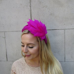 May include: A bright pink headband with a large, layered feather design. The headband is worn by a person with long blonde hair.