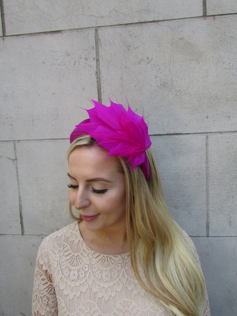 May include: A bright pink headband with a large, layered feather design. The headband is worn by a person with long blonde hair.