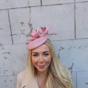 Può includere: Un fascinator rosa chiaro con base circolare e dettagli decorativi a fiocco e piume. Il copricapo è realizzato in materiale tessuto ed è progettato per essere indossato sulla testa. Un accessorio elegante per occasioni speciali.