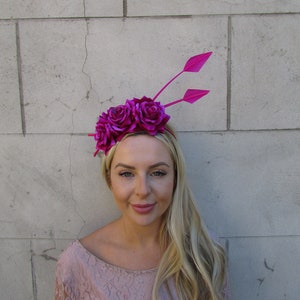 Pode incluir: Uma faixa de cabelo de flores rosa com duas penas. A faixa de cabelo é feita de flores de tecido e tem uma pena rosa de cada lado.