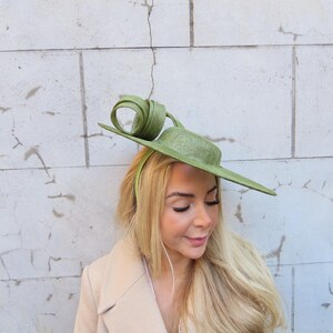 May include: A light green hat with a unique, asymmetrical design. The hat features a flat brim and a decorative swirl on top. The hat is worn with a light-colored coat. The hat is a stylish accessory.