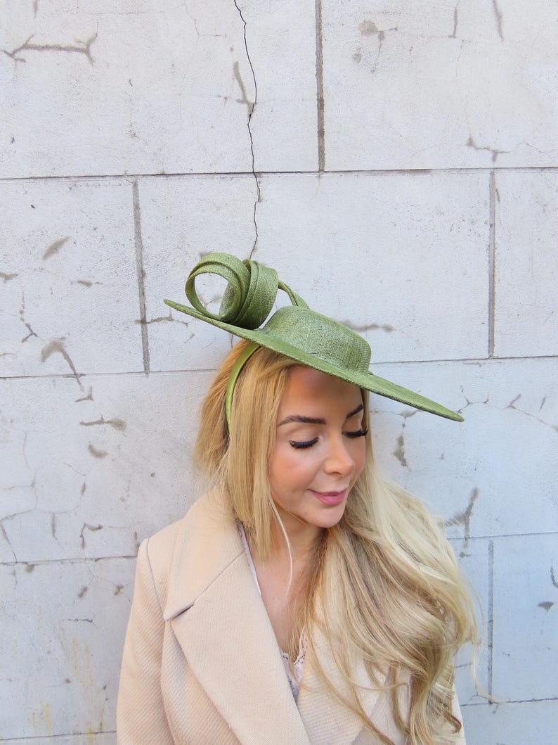 May include: A light green hat with a unique, asymmetrical design. The hat features a flat brim and a decorative swirl on top. The hat is worn with a light-colored coat. The hat is a stylish accessory.