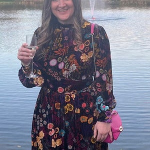 May include: A woman wearing a long, dark floral dress with a pink feathered headband. She is holding a glass of champagne and a small pink purse. She is standing in front of a lake with a fountain in the background.