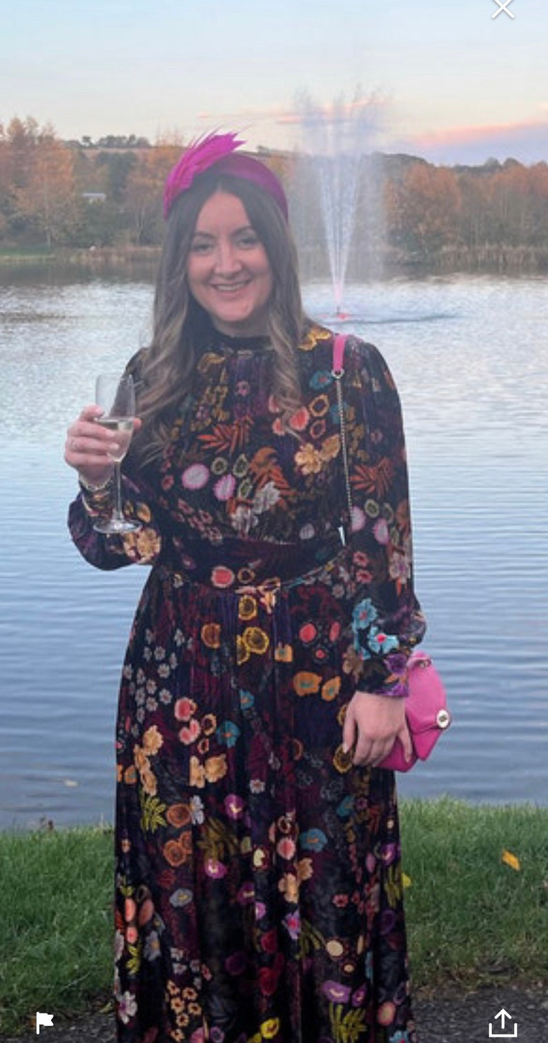 May include: A woman wearing a long, dark floral dress with a pink feathered headband. She is holding a glass of champagne and a small pink purse. She is standing in front of a lake with a fountain in the background.
