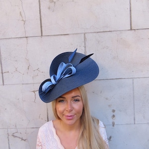 May include: A navy blue hat with a wide brim, adorned with a light blue ribbon and feathers. The hat has a structured design, suitable for formal events. The hat is a stylish accessory.