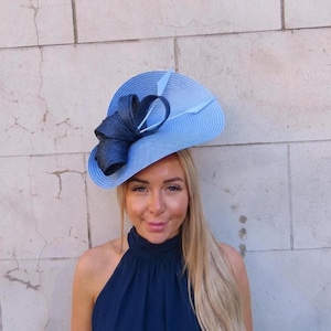 May include: A light blue fascinator hat featuring a wide, circular brim with a textured surface. The hat is adorned with a large, dark navy blue fabric bow detail and two thin, pointed quill-like accents extending from the center of the bow.