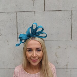 May include: A teal blue fabric headband with a large bow. The headband is worn by a person with long blonde hair.