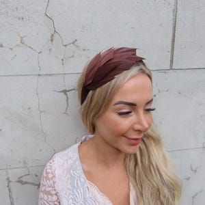 Könnte beinhalten: Ein braunes Feder-Haarband. Das Haarband besteht aus mehreren Federn, die fächerförmig angeordnet sind. Die Federn haben eine satte, dunkelbraune Farbe. Das Haarband wird auf dem Kopf getragen.