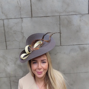 Puede incluir: Un sombrero gris con ala ancha, adornado con un lazo crema y plumas. El sombrero tiene forma circular y probablemente está hecho de material tejido. El sombrero es un accesorio de moda.