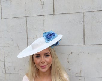 May include: A white hat with a wide brim, adorned with blue floral accents. The hat is worn by a person with long blonde hair and a light pink lace top. The background is a light-colored brick wall.