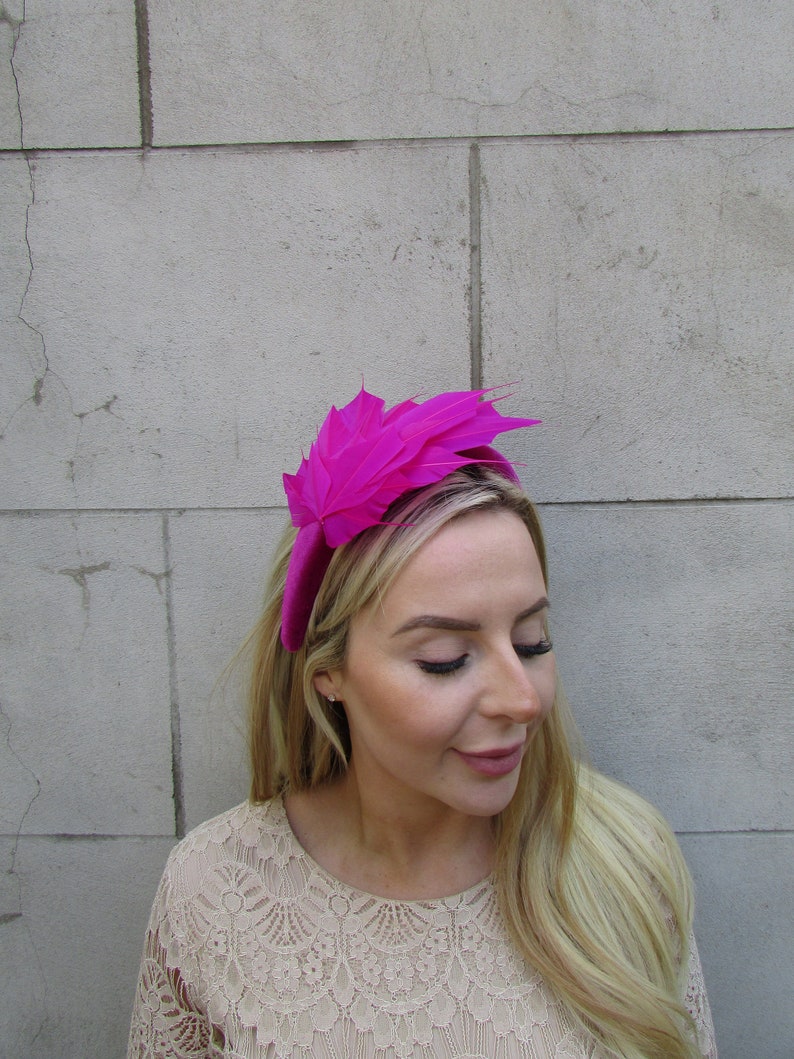 May include: A bright pink headband with a large feather accent. The headband is made of velvet and the feather is made of a soft, fluffy material.