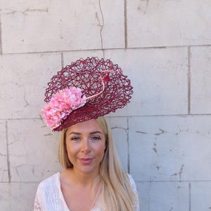 May include: A burgundy fascinator with a woven design and pink fabric flowers. The headpiece is worn by a person with blonde hair and a white lace top. The background is a light-coloured brick wall.