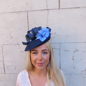 May include: A navy blue hat with a light blue flower and ribbon detail. The hat has a small brim and is likely for a special occasion. The hat is set against a light-colored, textured wall.